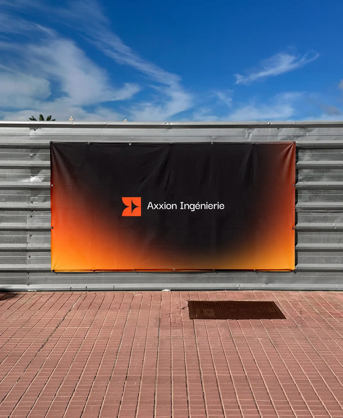 Une grande bâche noire à dégradé orange portant le logo Axxion Ingénierie, tendue sur une palissade de chantier, sous un ciel bleu.