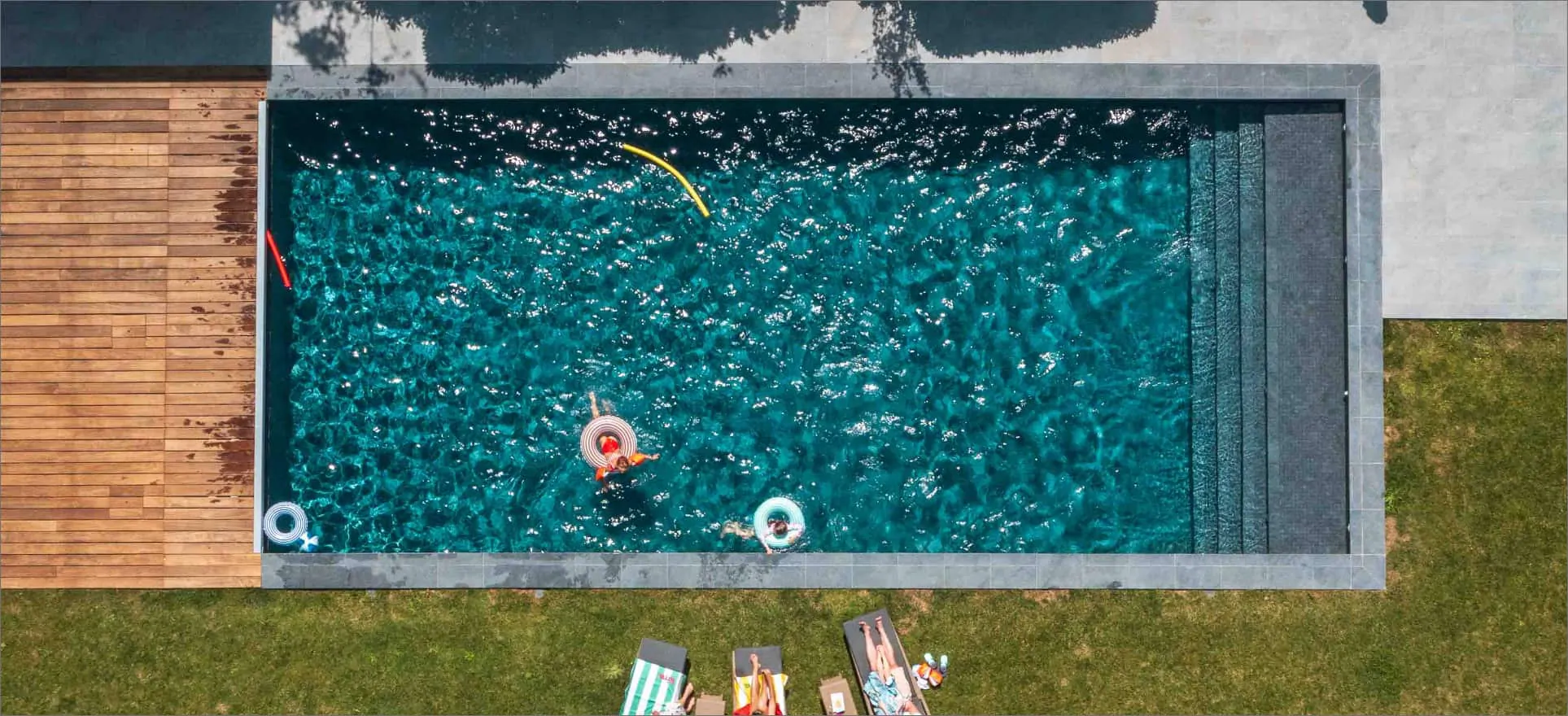 Une piscine rectangulaire vue du ciel, bordée d’une terrasse en bois et d’une pelouse ; une personne sur une bouée, des transats au bord.