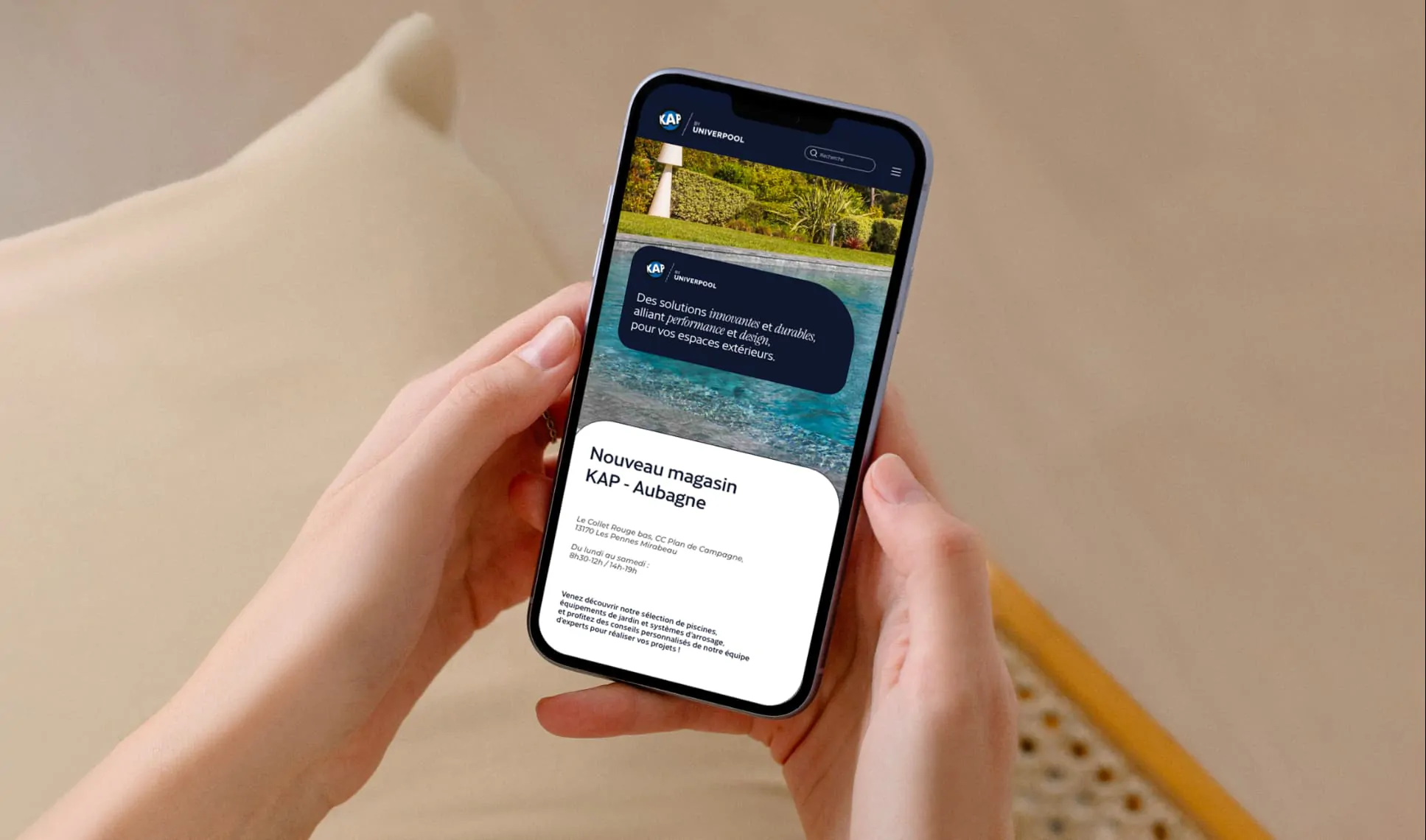 Deux mains tiennent un téléphone affichant le site de KAP by Univerpool, au-dessus d’un coussin beige.
