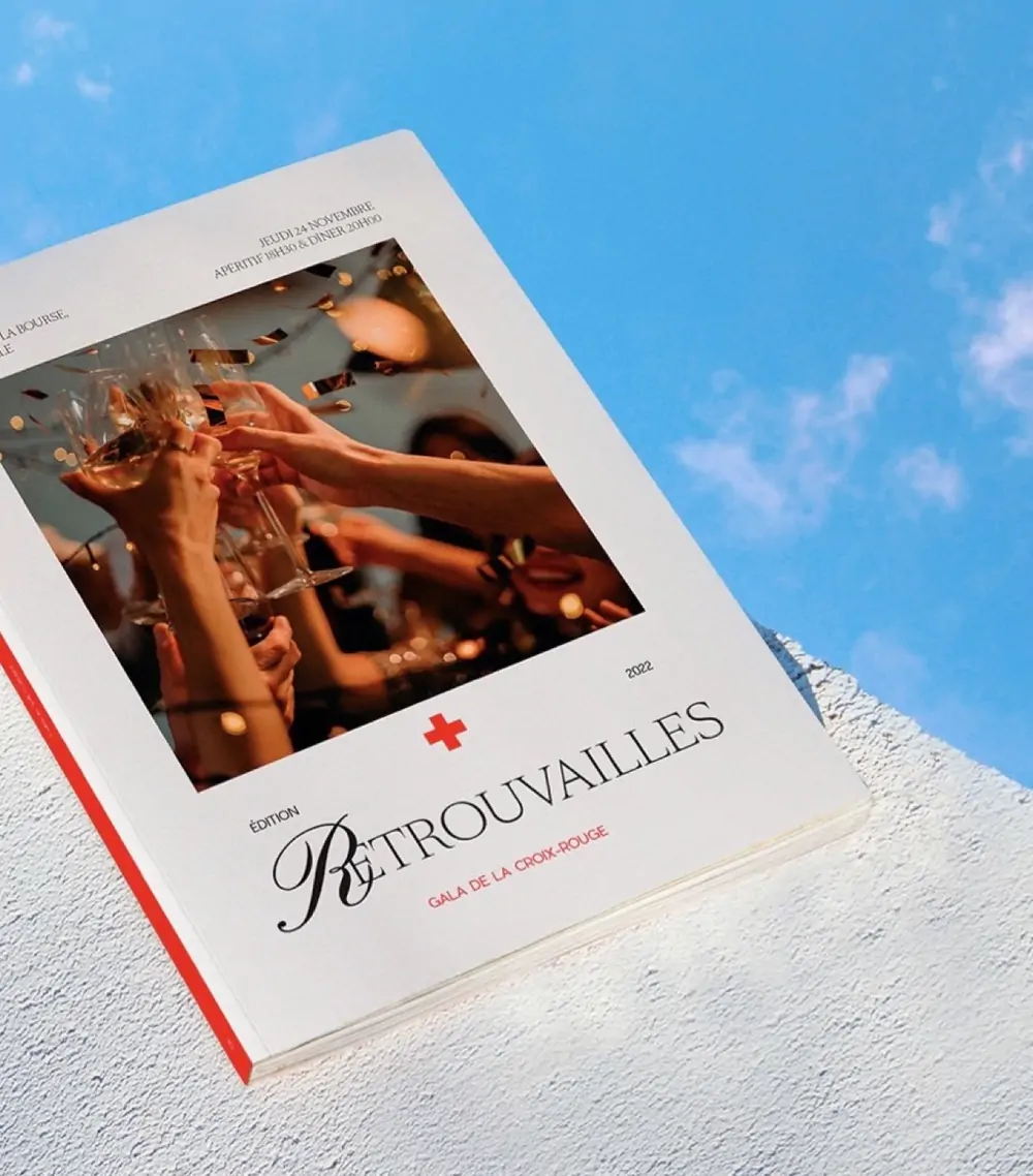 Gala de la Croix-Rouge — le catalogue « Retrouvailles », une couverture blanche à bandeau rouge, posé sur un ciel bleu